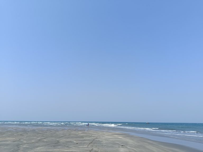 Beach with clear blue sky, gentle waves, and distant figures under the vastness of an open horizon.