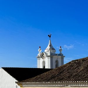 Tiled rooftops and a church bell tower, clock and cockerel weather vien against a cloudless vivid blue sky in Estói, Portugal
