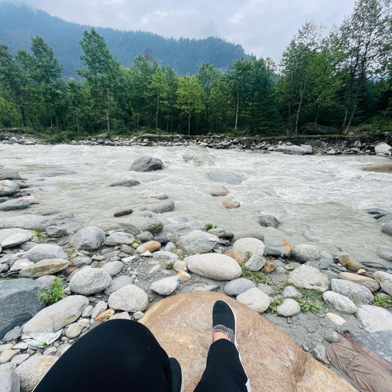 Feet and legs of the photographer on rocks beside a fast-moving stream. There are trees and hills in the background.