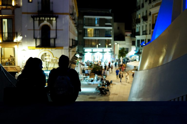 Back view of couple sitting on top of stairs at night, and a busy street ahead of them.