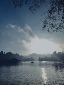 View of the sunrise in a misty morning in Kodaikanal with silhouette of people in the boat, trees and a hut in the frame.