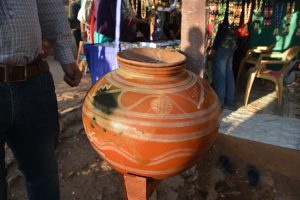 Decorative Matka or Matkia on its stand, a drinking water pot used to keep water cool in Indian villages. Market stalls clothing, beaded jewellery and two partial people can be seen.