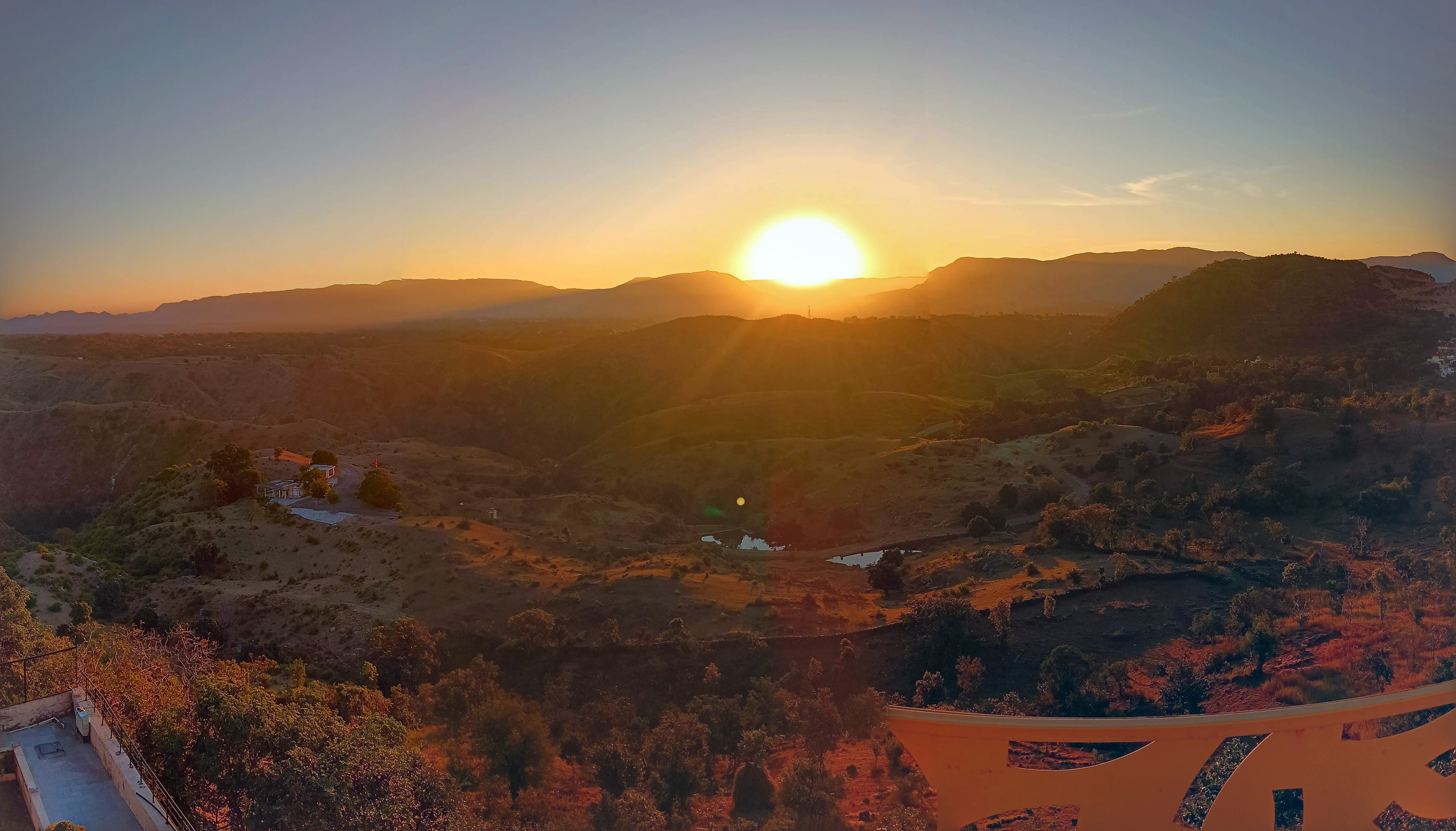 A panoramic view unveils a sunrise over a mountainous region. The sky transitions from a warm orange near the horizon to soft blue above. Sparse vegetation, undulating hills, and a glinting pond compose the serene landscape, with a lone building perched atop. Paths ribbon through the terrain.