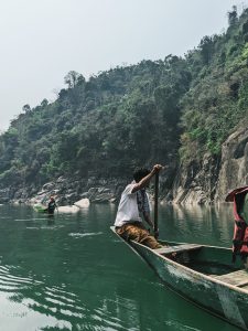 A man rowing a boat through the sea with an island with rocks and trees in the background. 