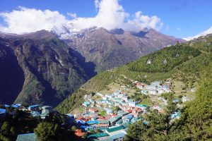 Beautiful Namche Bazaar situated on the side of a mountain with trees, and mountain ranges covered in snow in the background. 