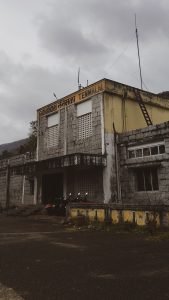 Thenmala Railway Station, motorcycles parked , a wooden ladder on the first floor reaches up to the roof. Litter lies on the road, stormy sky overhead.