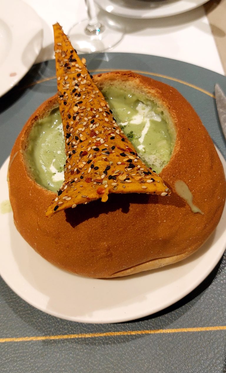 Creamy soup served in a bread bowl with a tall, seed-encrusted breadstick on a white plate.