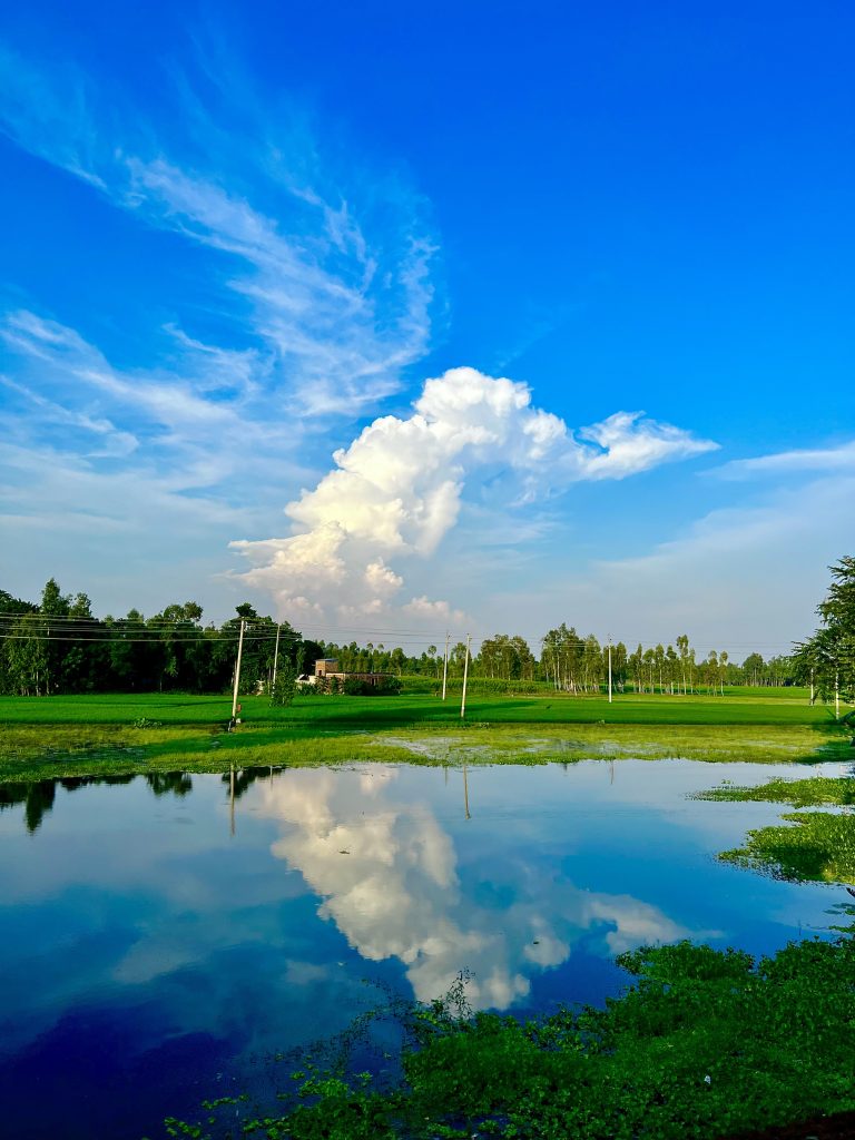 A vibrant landscape with a large cumulus cloud reflected in a serene lake surrounded by greenery under a blue sky with wispy clouds.