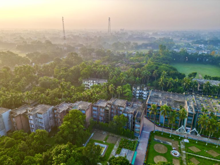 Drone landscape of a polytechnic institute featuring several buildings surrounded by different kinds of trees. In the background distance are more buildings and two metal towers with a yellow/orange sky.