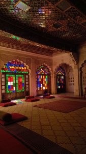 A hall with a beautiful coloured tinted glass windows, carpet and cushions on the floor, and glasses on the roof reflecting the colors.