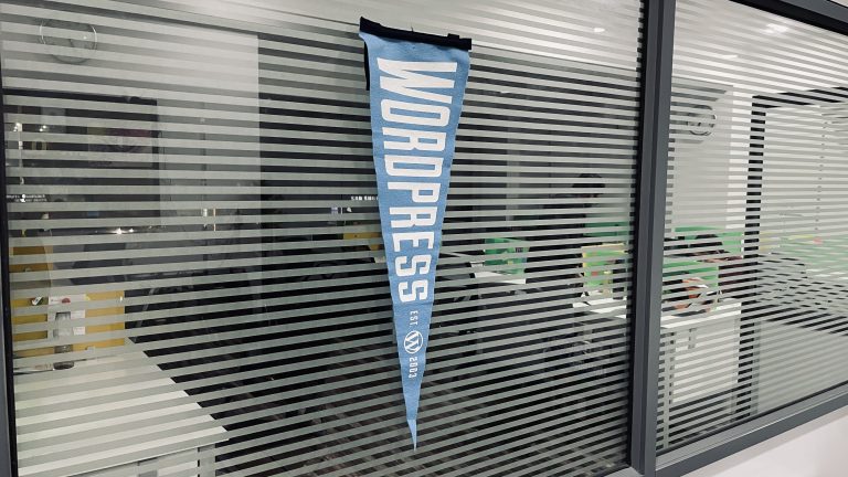 A WordPress pennant hangs in a modern office setting, its bold logo on display against the geometric patterns of frosted glass partitions, reflecting a creative and tech-savvy environment.