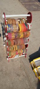 Colourful beaded and metallic bangle bracelets hang ready for sale from a stand on a pavement in the sunshine.  