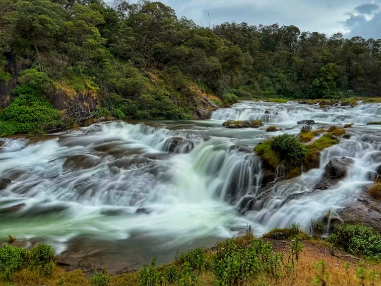 Long exposure photo of a waterfall in the middle of a lush green forest with a cloudy sky.