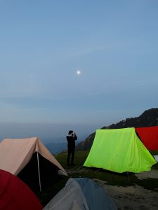 A man standing besides colourful tents on a mountain taking picture of the valley. 