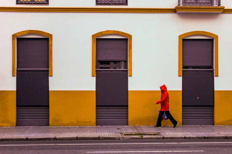 Person on a red raincoat with hood, passing in front of a white and yellow building