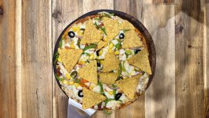 Pizza with corn, tortilla chips, olives, and peppers on a wooden table
