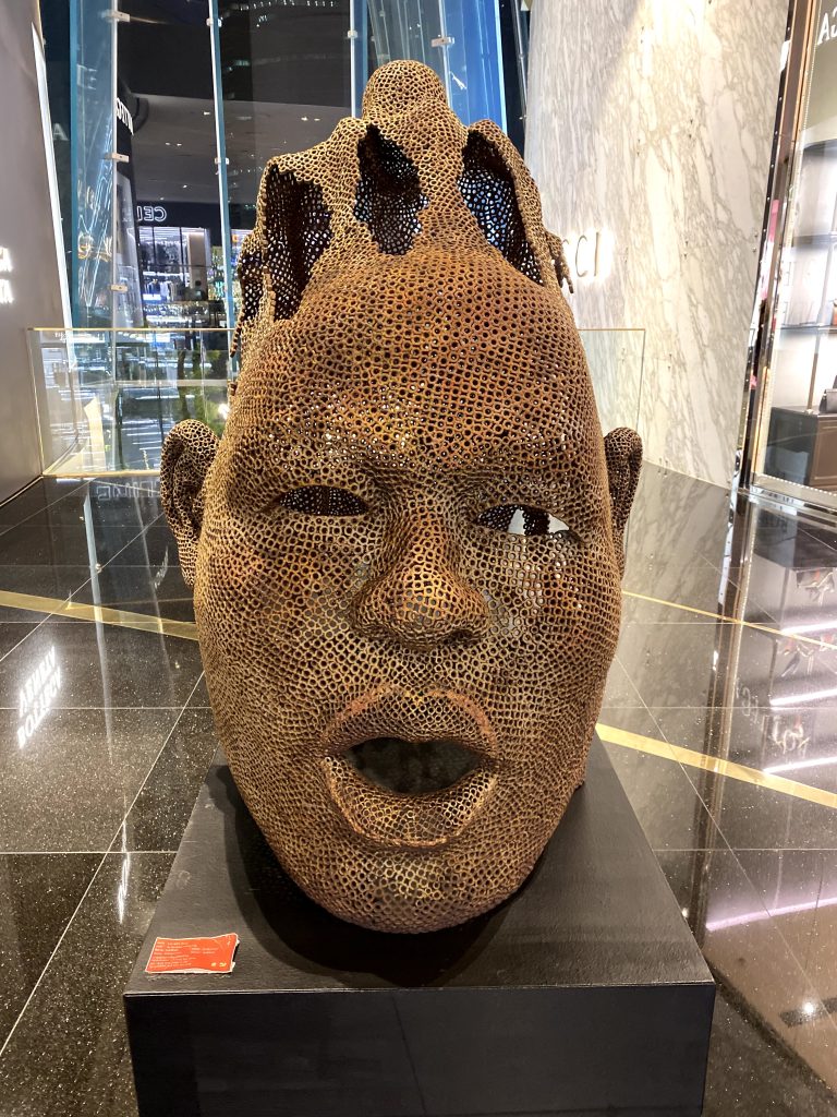 A metal sculpture of a human face with open head and mouth on top of a pedestal