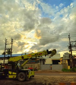 A yellow mobile crane on the side of a road with power lines and utility poles against a backdrop of a dramatic sky with clouds during sunset.