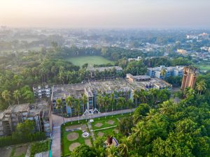 Ariel vista of a polytechnic institute surrounded by grass and palm trees on a hazy day, a sizable settlement beyond it.