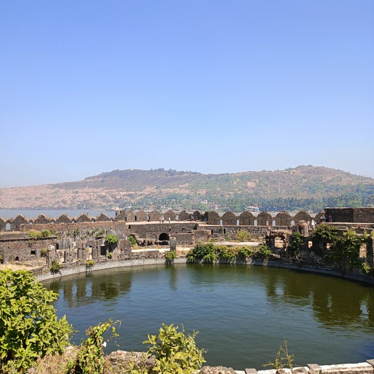 Inside the walls of the Murud-Janjira fort, including a large, deep pool of water. Hills and the sea are in the background.