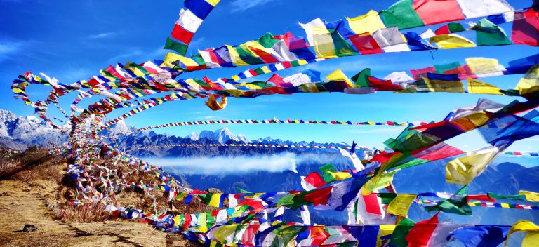 Brightly colored prayer flags dance in the wind above Amayangri and the Himalayas, set against a backdrop of a vivid blue sky.