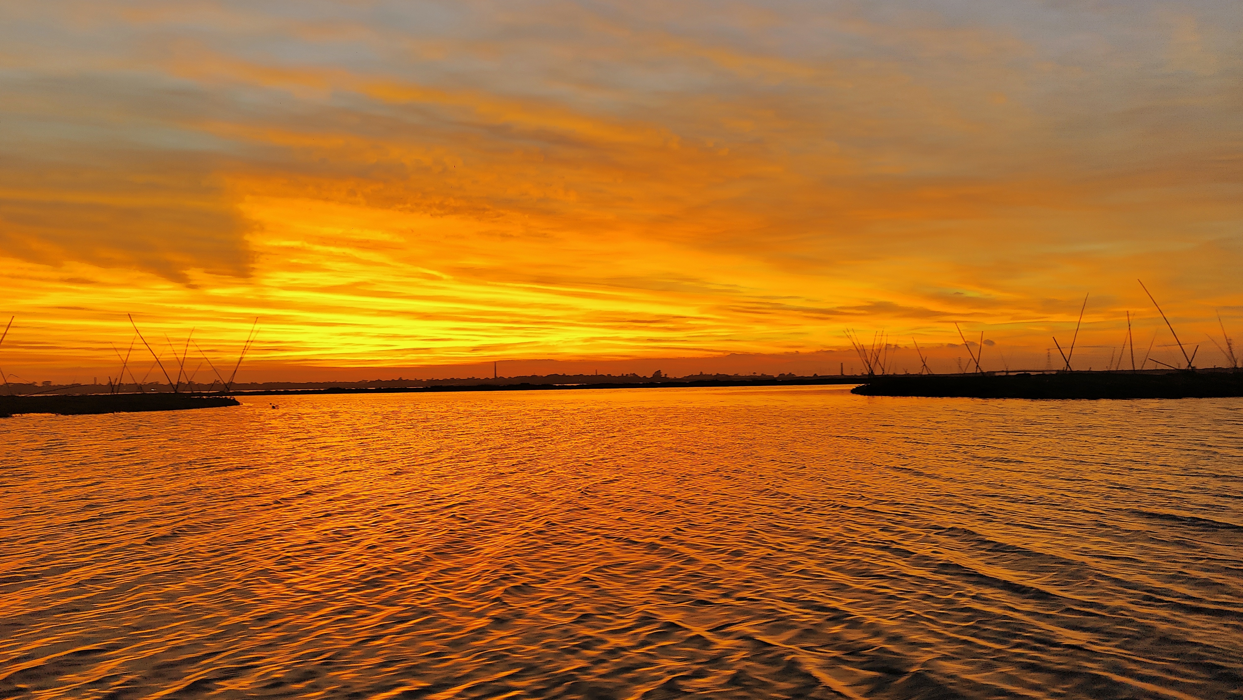 Titas River at sunset the entire vista bathed in bright orange sunlight, buildings and tall poles in silhouette. 