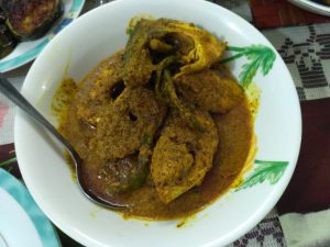 A plate of spicy Indian food with what appears to be a rich, thick sauce, accompanied by a metal spoon, placed on a table with a patterned tablecloth.