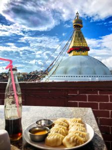 Boudhanath Stupa is adorned with prayer flags, accompanied by momos and chilled beverages in the foreground. 