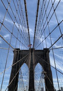 布鲁克林大桥塔的吊索视图，在明亮的蓝天，美国的星条旗和斯蒂普斯旗在桥上飘扬，飞机在桥上飞过。
