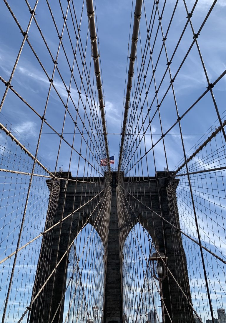 Suspension cable view of Brooklyn Bridge Tower, on bright blue sky day, Star and Stipes flag of the USA flies on the bridge as an aircraft flies overhead.