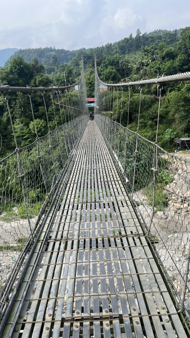 Small hanging metal bridge over an empty river with lush greenery in the background.