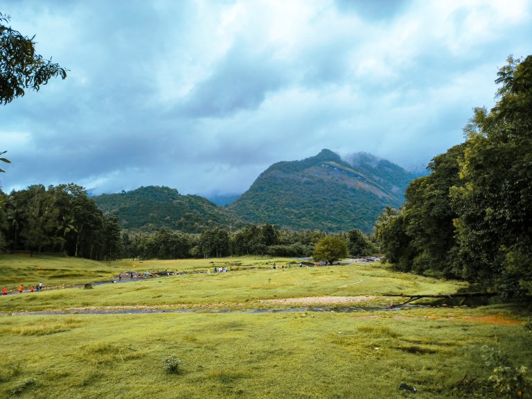 People walk along a stream in the distance through an open field with mountains in the background