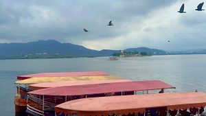 View larger photo: A serene lake with boats gently floating on its surface, surrounded by birds flying in the sky overhead.