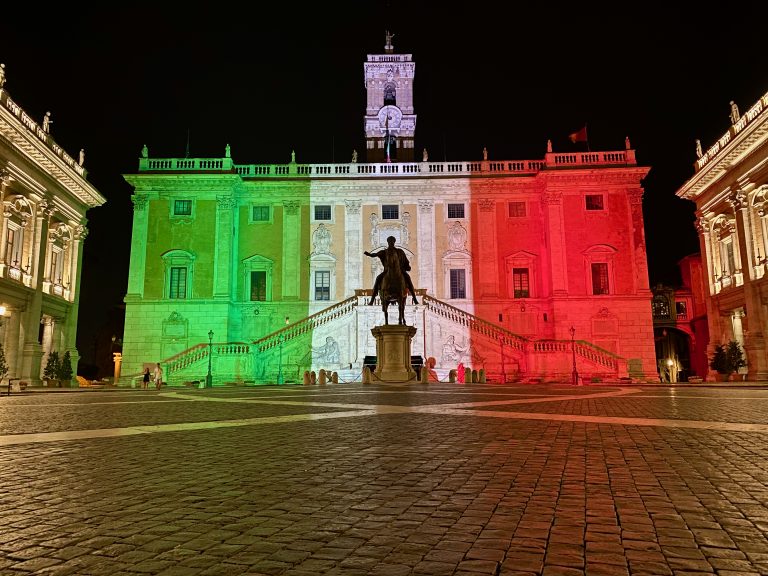 The Senatorial palace on the Capitoline Hill in Rome, Italy at night. The palace is illuminated in the colors of the Italian flag – green, white, red. In front of the palace is a bronze statue.