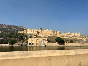 Looking at Amber Fort across the water (Front View). It's a fortress/palace with many walls and turrets built on a hill.