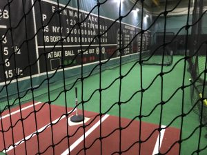 A batting tee on home plate inside a batting cage. The wall is painted with a replica of the Fenway Park scoreboard. An L screen is setup at pitching distance.
