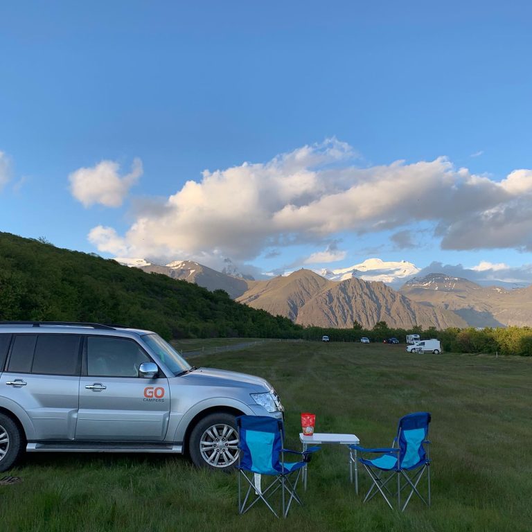 Iceland chill zone: Car parked on a lawn with chairs and a desk besides it with a beautiful view of mountain ranges in the background.