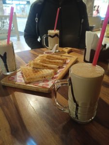 Three glasses of milkshake served with sandwiches on a wooden table.