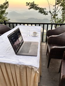 Embracing the Remote Office with a View to Inspire. A laptop, cup of coffee and a glass of water placed on table covered with white cloth with a beautiful view of mountain range in the background. 