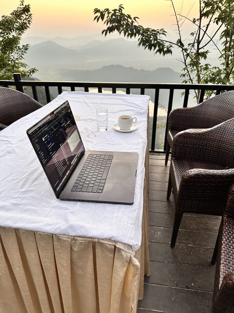 Embracing the Remote Office with a View to Inspire. A laptop, cup of coffee and a glass of water placed on table covered with white cloth with a beautiful view of mountain range in the background.