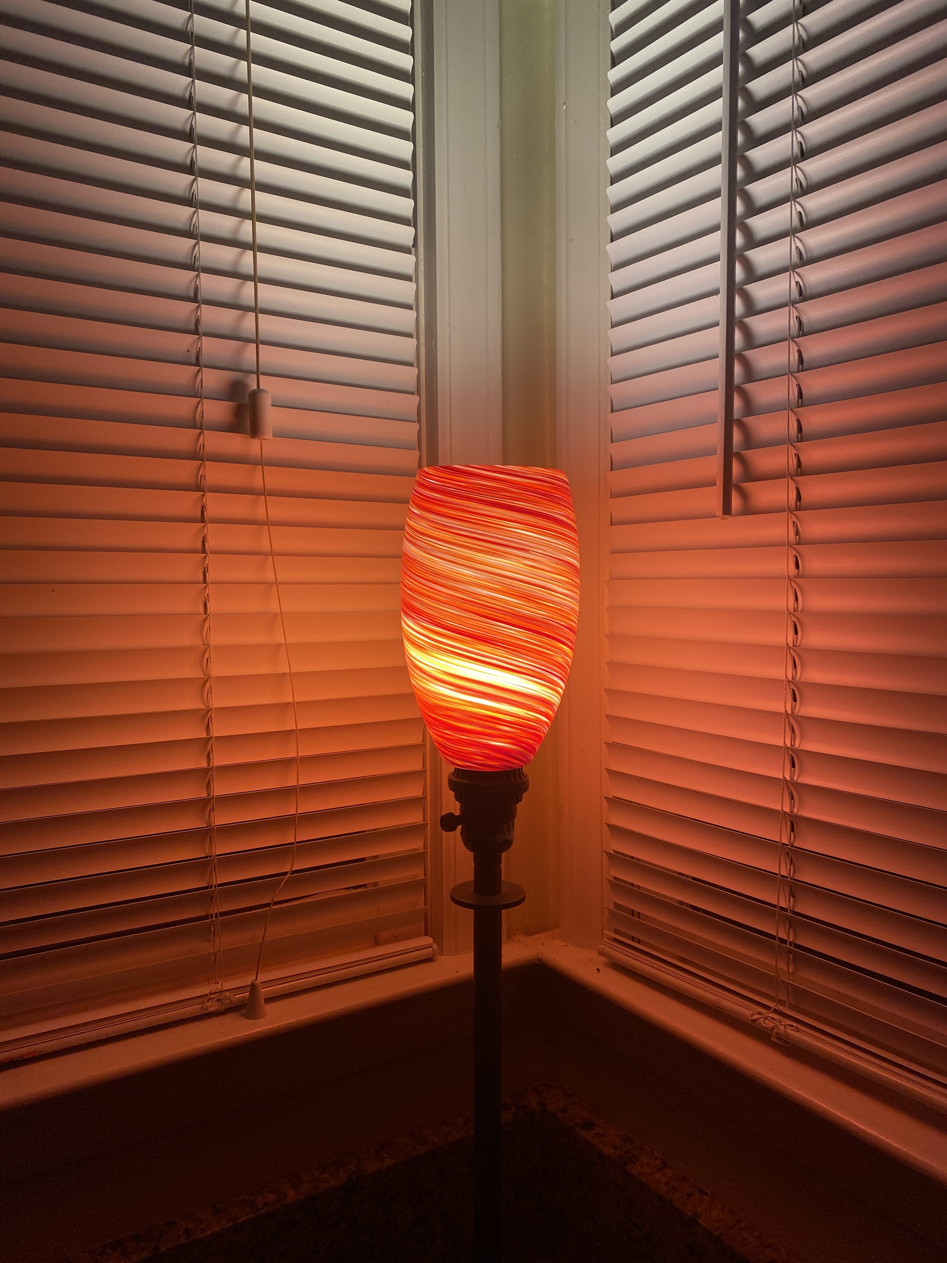 red swirled glass table top light fixture in the corner of two windows with the blinds closed