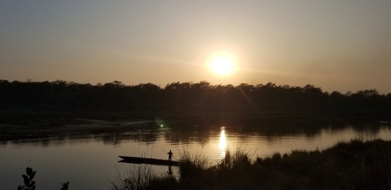 Sun is setting over a lake. A person is rowing a boat near the shore.