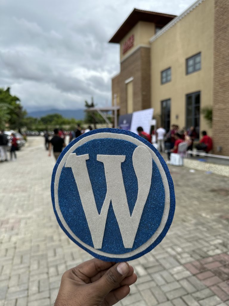 A hand holding a WordPress handmade logo with people and a buinding behind.