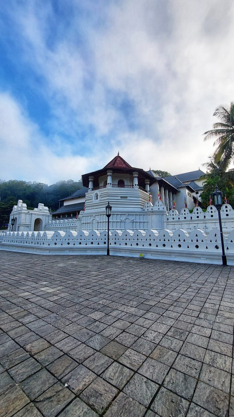 The Paththirippuwa, also known as the octagonal pavilion temple which houses the sacred tooth relic of budha. It is world heritage site declared by UNESCO, within the Temple of the Sacred Tooth Relic in Kandy, Sri Lanka. It’s a white building surrounded by trees and a blue sky with wispy clouds.