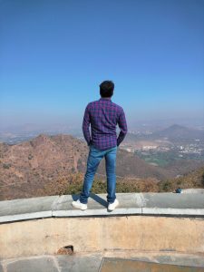 A man wearing plaid shirt and  blue jeans standing facing mountains under a clear blue sky.
