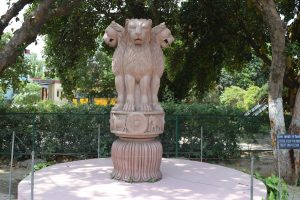 Statue depicting the National Emblem of India in Sarnath, surrounded by trees and fenced off. A bus passing by and buildings beyond.