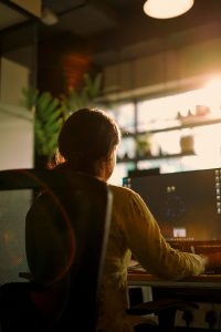 A girl working on a computer system near a window with sunlight is shining through it. 