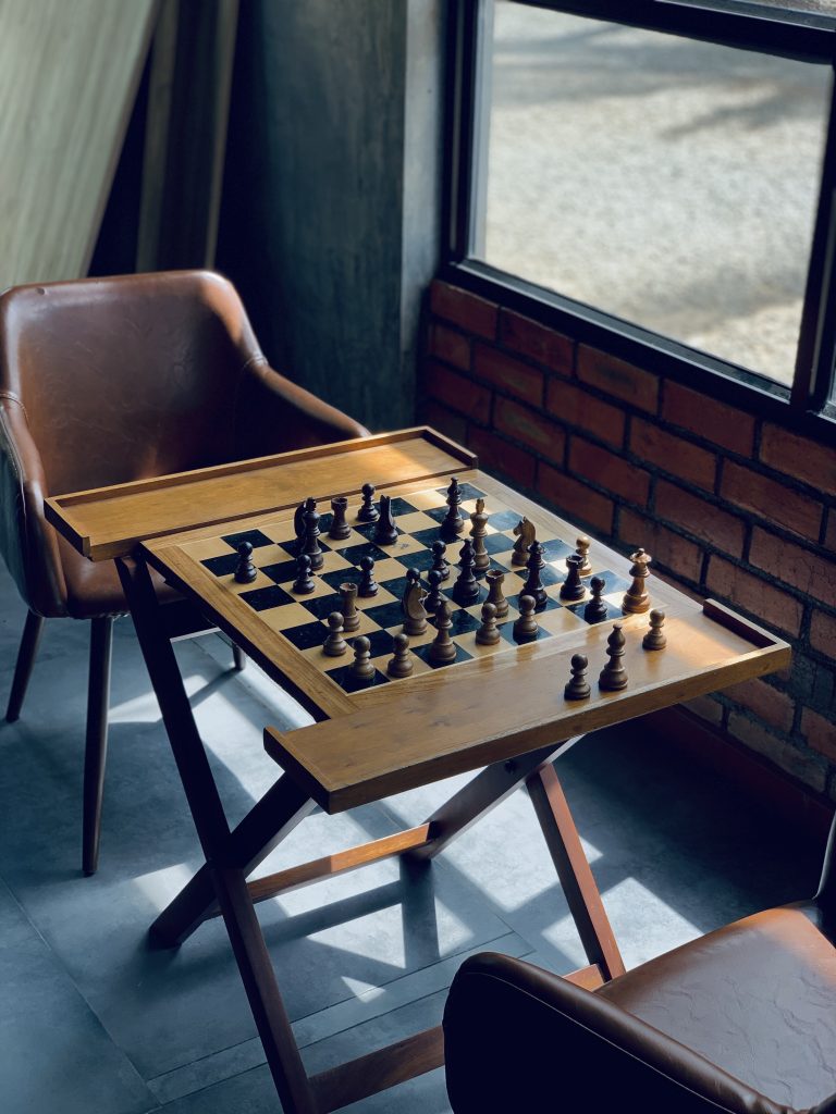 chess Board on a table with chairs in the side of a window.