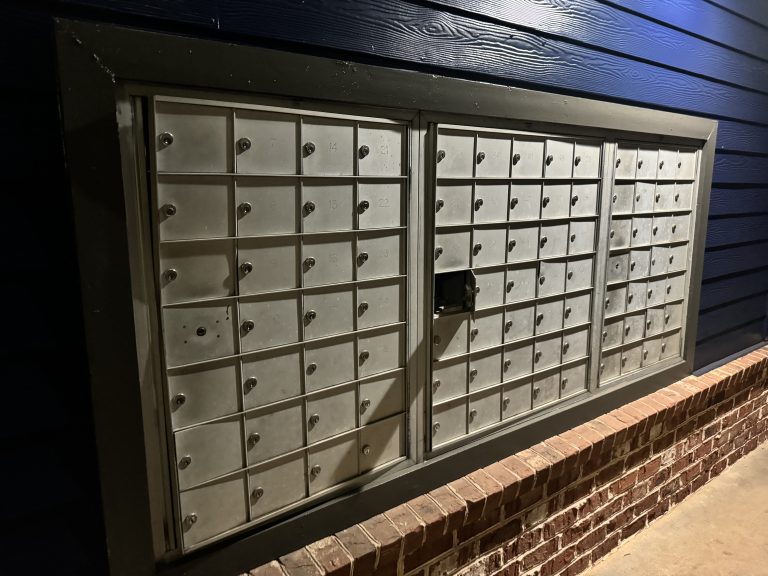 Metal lockable post boxes in three blocks of four by seven, fitted into a wooden painted wall, above a brick wall. The wood grain is clearly visible.