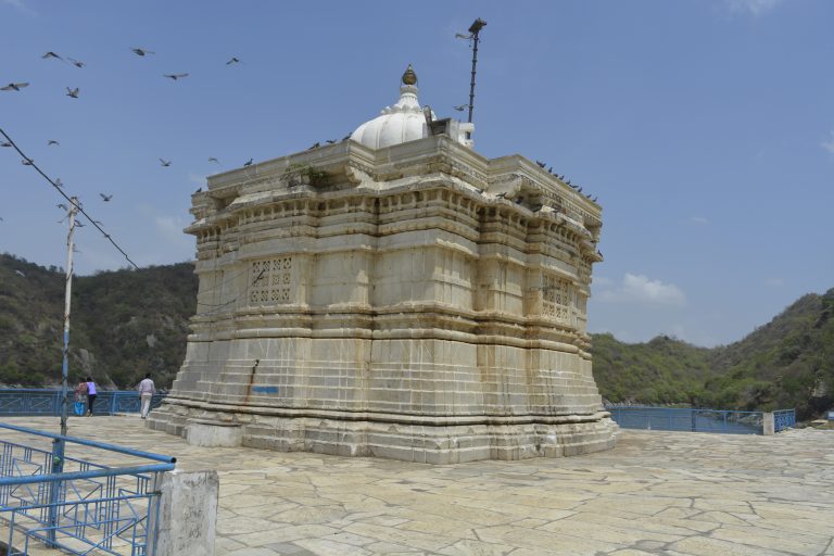 A white stone monument with a blue sky and birds flying overhead near Jaisamand lake in Udaipur, India